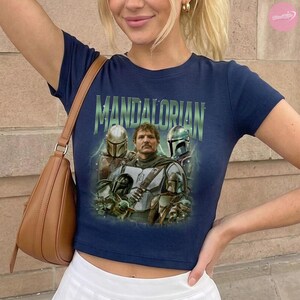 May include: Navy blue crop top featuring a graphic of characters from the Mandalorian series. The word "MANDALORIAN" is printed in green across the top. The image includes characters in helmets and armor.