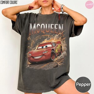 May include: Pepper-colored t-shirt featuring a graphic of Lightning McQueen from the movie Cars. The design includes the name "MCQUEEN" in a distressed font above the car, with lightning bolts and a speedometer design.