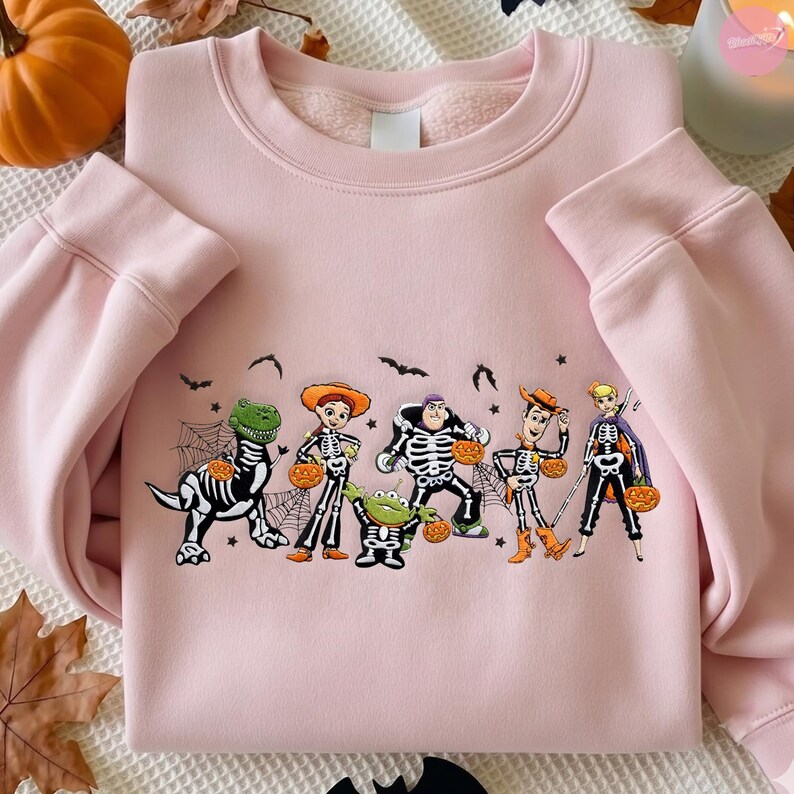 May include: A light pink crewneck sweatshirt featuring a graphic print of six animated characters dressed in skeleton costumes for Halloween. The characters are holding orange jack-o'-lanterns, surrounded by spiderwebs and small black bats.