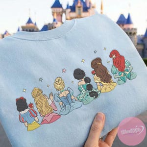 May include: Light blue sweatshirt featuring an embroidered design of six cartoon princesses. The princesses are depicted from the back, with detailed hairstyles and colorful dresses. Gold stars are scattered around the design. The sweatshirt is held up by a hand.