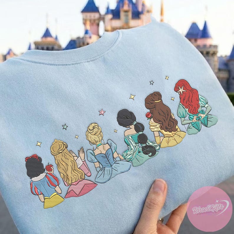 May include: Light blue sweatshirt featuring an embroidered design of six cartoon princesses. The princesses are depicted from the back, with detailed hairstyles and colorful dresses. Gold stars are scattered around the design. The sweatshirt is held up by a hand.