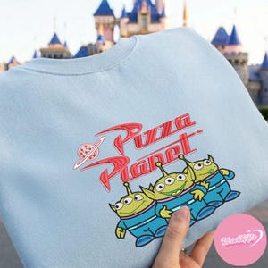 May include: Light blue sweatshirt featuring the Pizza Planet logo in red and a cartoon image of three green aliens in blue uniforms. The sweatshirt is a crew neck style.