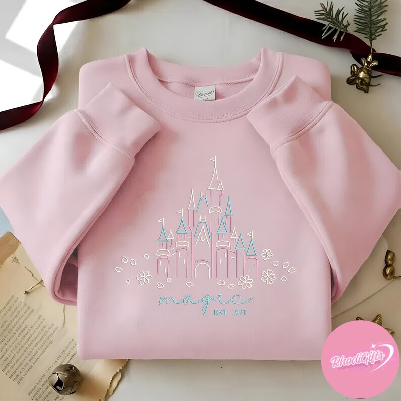 May include: A light pink sweatshirt with a castle graphic in pink and teal. The word "magic" is written in teal script below the castle, with "EST 1971" underneath. White floral accents surround the text and castle.