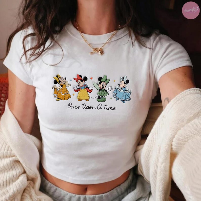 K&ouml;nnte beinhalten: Wei&szlig;es Crop-Top mit vier Cartoon-Figuren in farbenfrohen Kleidern. Der Text "Once Upon a Time" ist unter den Figuren aufgedruckt. Das Shirt hat kurze &Auml;rmel und einen Rundhalsausschnitt.