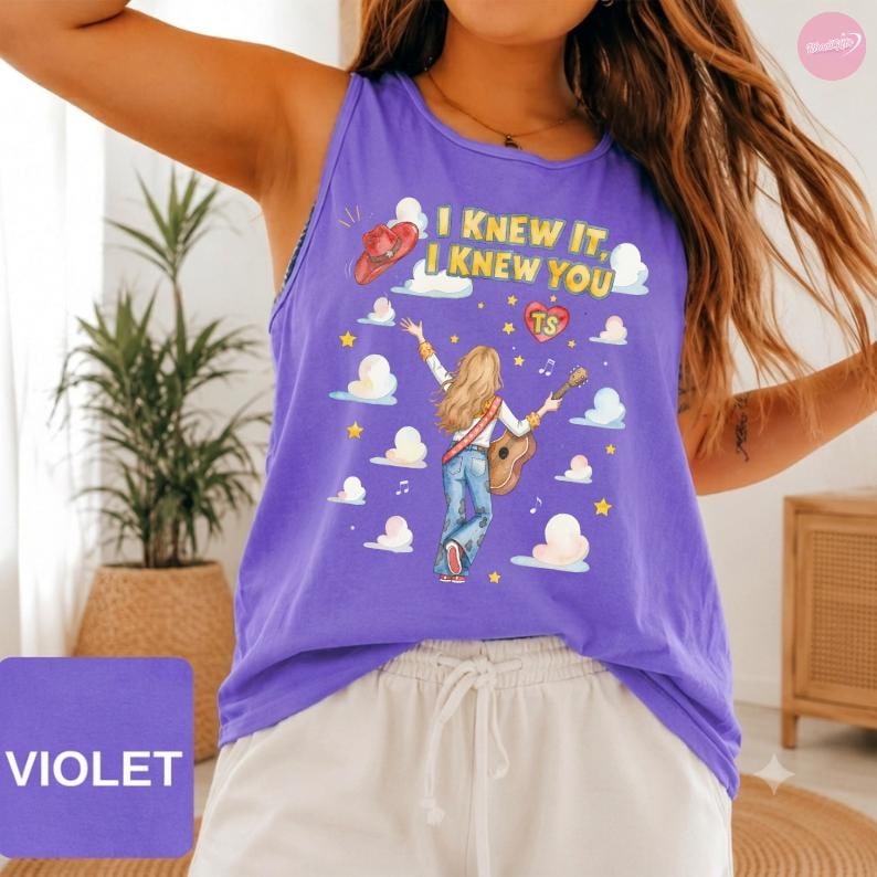 May include: A violet tank top featuring a graphic of a person with long blonde hair playing an acoustic guitar, surrounded by clouds, stars, and musical notes. The text reads I KNEW IT, I KNEW YOU and TS inside a heart, with a red cowboy hat.