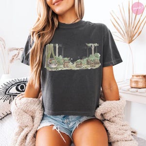 Puede incluir: Camiseta gris oscuro con una escena de bosque con varios personajes de Grogu. El dise&ntilde;o incluye a Grogu en una c&aacute;psula flotante y otras figuras de Grogu entre los &aacute;rboles. La palabra "Cool" es visible. La camiseta tiene mangas cortas y cuello redondo.