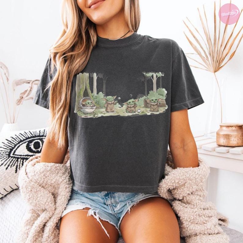 Puede incluir: Camiseta gris oscuro con una escena de bosque con varios personajes de Grogu. El dise&ntilde;o incluye a Grogu en una c&aacute;psula flotante y otras figuras de Grogu entre los &aacute;rboles. La palabra "Cool" es visible. La camiseta tiene mangas cortas y cuello redondo.