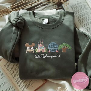 Peut inclure: Sweat-shirt vert olive avec broderie Walt Disney World. Le motif comprend Mickey Mouse, le ch&acirc;teau de Cendrillon, Spaceship Earth et un arbre. Les mots "Walt Disney World" sont brod&eacute;s en dessous.