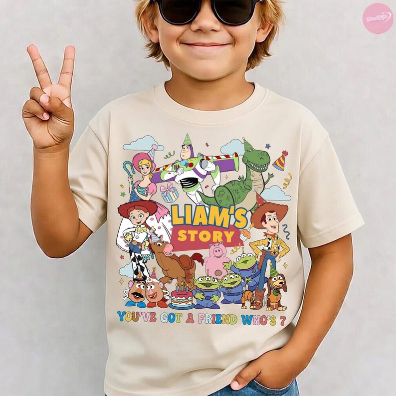 May include: Beige short-sleeved t-shirt featuring a colorful graphic of characters from the movie Toy Story. The graphic includes the text "LIAM'S STORY" and "YOU'VE GOT A FRIEND WHO'S 7?" with various cartoon characters.