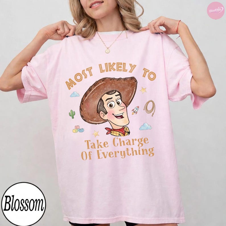 Op de afbeelding: Een lichtroze t-shirt met een afbeelding van een cartooncowboy met een bruine hoed. De tekst luidt Most Likely To Take Charge Of Everything. Kleine illustraties van een cactus, raket, lasso en bouwblokken omringen het personage.