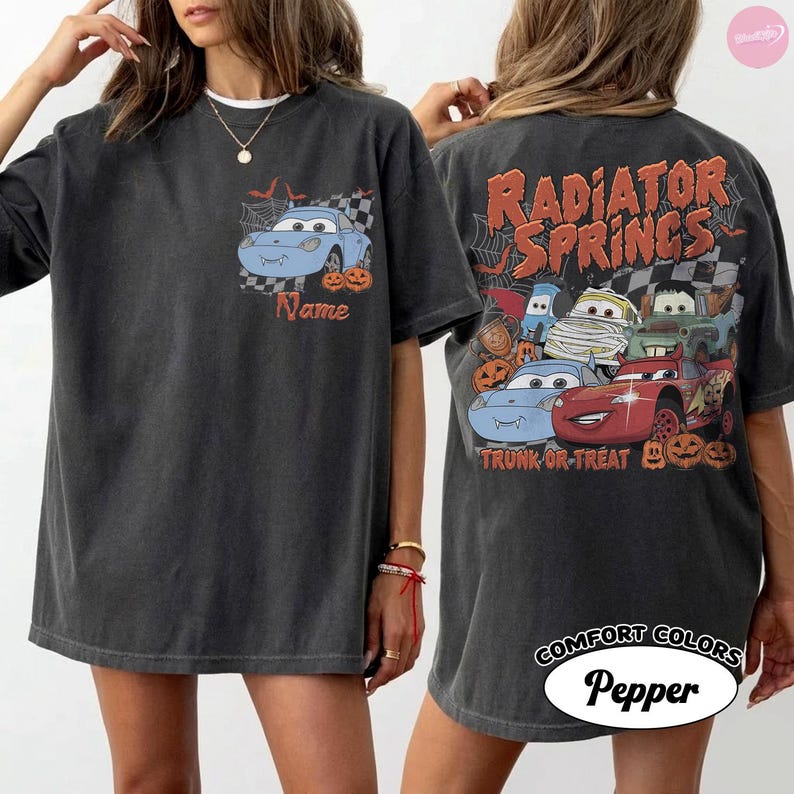 May include: A charcoal grey t-shirt featuring a small graphic on the front left chest of a blue car with vampire fangs and pumpkins. The back displays a large graphic of multiple cars, pumpkins, and the text Radiator Springs Trunk or Treat.