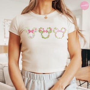 May include: Cream-colored, short-sleeved t-shirt featuring three Mickey Mouse head designs. Each design is a floral arrangement with pink bows and flowers. The shirt has a round neckline and is made of a soft, comfortable material.