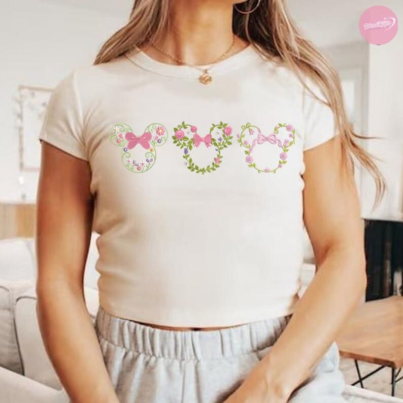 May include: Cream-colored, short-sleeved t-shirt featuring three Mickey Mouse head designs. Each design is a floral arrangement with pink bows and flowers. The shirt has a round neckline and is made of a soft, comfortable material.