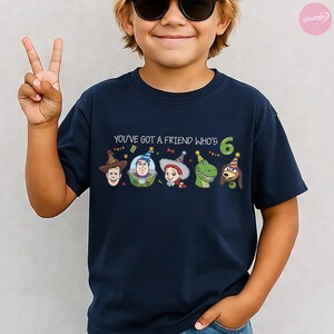K&ouml;nnte beinhalten: Marineblaues T-Shirt mit dem Text "YOU'VE GOT A FRIEND WHO'S 6" und Cartoon-Charakteren. Die Grafiken zeigen Woody, Buzz Lightyear, Jessie, Rex und Slinky Dog. Die Zahl 6 ist gr&uuml;n.