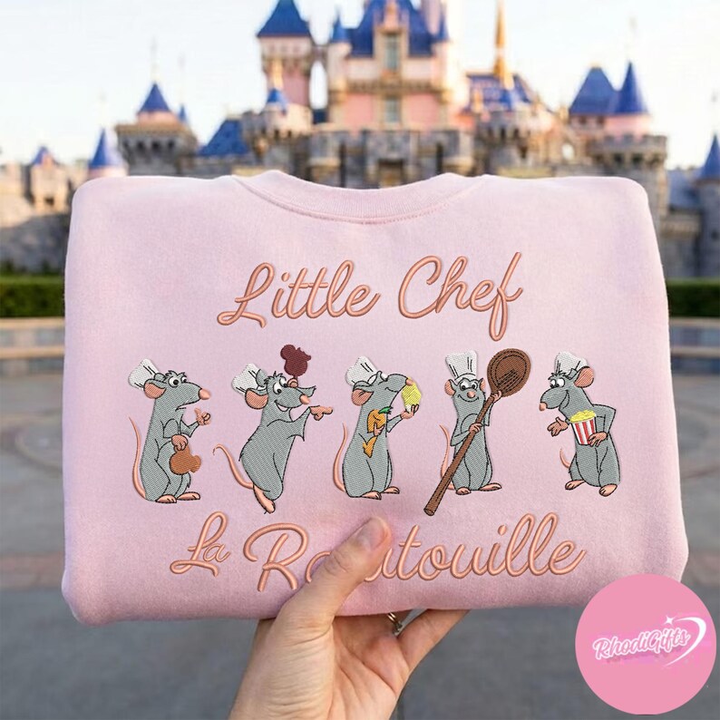 Pode incluir: Camisola rosa claro com bordados de ratos de desenho animado usando chap&eacute;us de chef. As palavras "Little Chef" e "La Ratatouille" s&atilde;o bordadas em uma cor correspondente. A camisola &eacute; de gola redonda.
