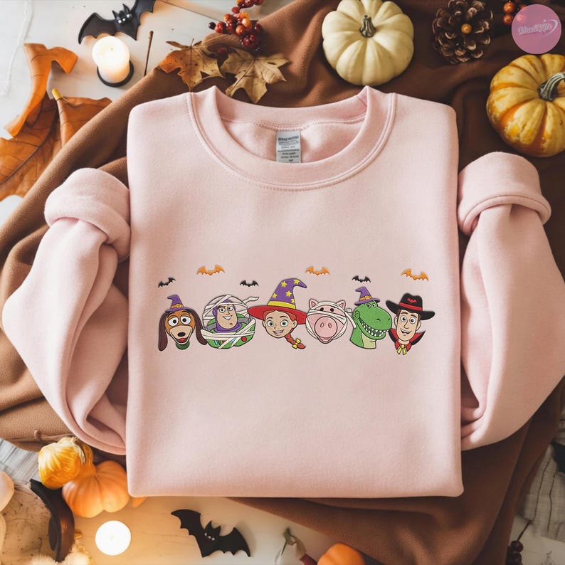 May include: A light pink crewneck sweatshirt featuring a Halloween-themed graphic of Toy Story characters in costumes. The design includes Slinky Dog, Buzz Lightyear, Jessie, Hamm, Rex, and Woody. Small bats and pumpkins are also included in the design.