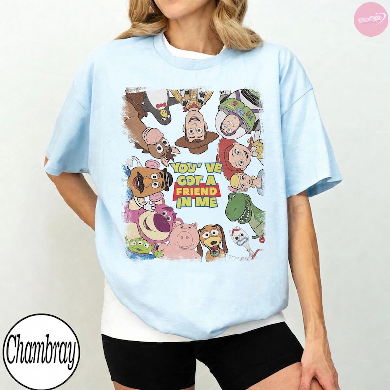 May include: Light blue t-shirt featuring a colorful graphic of characters from the animated film Toy Story. The graphic includes Woody, Buzz Lightyear, Jessie, and others, with the text "You've Got a Friend in Me".