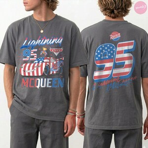 May include: Gray t-shirt featuring a graphic of Lightning McQueen from the movie Cars. The front design includes the text "Lightning 95 McQueen" with an American flag theme. The back of the shirt displays "95" with a star-spangled banner design and the text "Lightning McQueen".