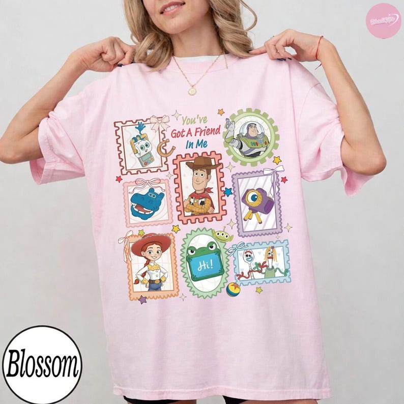 May include: A light pink t-shirt featuring a graphic print of various Toy Story characters inside postage stamp-style frames. The design includes Buzz Lightyear, Woody, Jessie, and other figures with the text You've Got A Friend In Me.