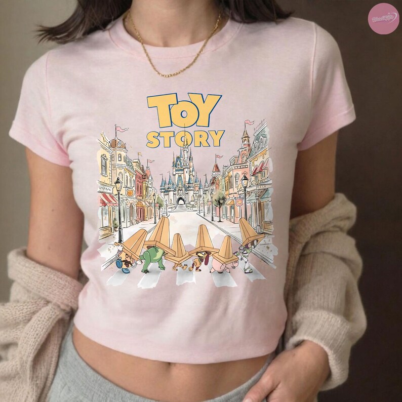 May include: A light pink t-shirt featuring the words "TOY STORY" in yellow block letters above a colorful illustration. The illustration depicts a street scene with cartoon characters crossing a crosswalk.