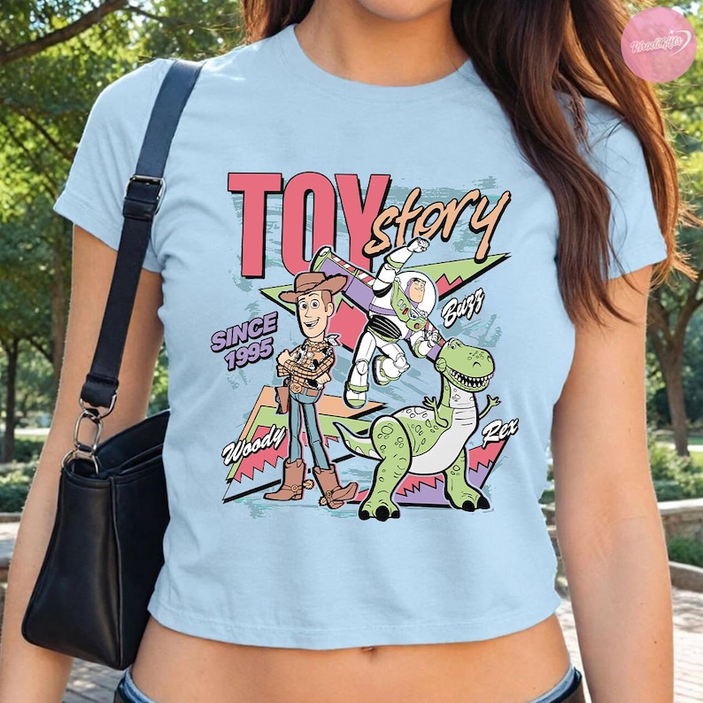 Op de afbeelding: Lichtblauw T-shirt met de tekst "Toy Story" in roze en geel, met Woody, Buzz Lightyear en Rex de dinosaurus. Het shirt heeft ook de tekst "Since 1995".