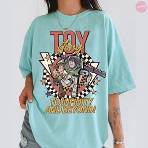 May include: A pale blue t-shirt with a graphic of Woody and Buzz Lightyear from Toy Story, with the words "TOY STORY" in red and yellow. The phrase "TO INFINITY AND BEYOND!" is printed below the characters, with a checkered background and star accents.