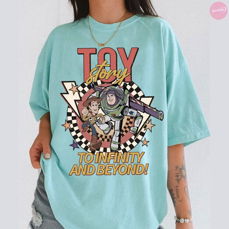 May include: A pale blue t-shirt with a graphic of Woody and Buzz Lightyear from Toy Story, with the words "TOY STORY" in red and yellow. The phrase "TO INFINITY AND BEYOND!" is printed below the characters, with a checkered background and star accents.