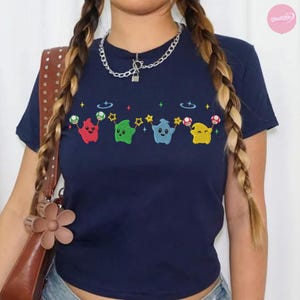 Puede incluir: Camiseta azul marino con un dise&ntilde;o bordado de cuatro fantasmas de dibujos animados en rojo, verde, azul y amarillo, con estrellas y setas. La camiseta tiene mangas cortas y cuello redondo.