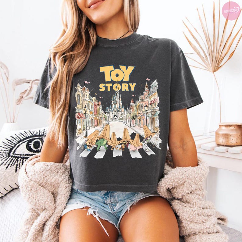 May include: Dark gray t-shirt with the words "TOY STORY" in yellow. The graphic depicts a cartoon scene of characters crossing a crosswalk in front of a castle and buildings. The shirt is a crew neck.