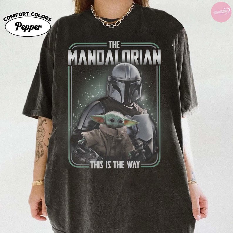 K&ouml;nnte beinhalten: Dunkelgraues T-Shirt mit einer Grafik von The Mandalorian und Grogu. Das Design enth&auml;lt den Text "THE MANDALORIAN" und "THIS IS THE WAY" in Wei&szlig;. Das Shirt ist von der Marke Comfort Colors.