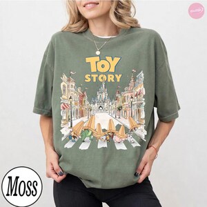 May include: Olive green t-shirt featuring the words "Toy Story" in yellow, with a graphic of the characters crossing a street in front of a castle. The shirt has a crew neck and short sleeves.