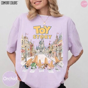 May include: Orchid-colored Comfort Colors t-shirt featuring a graphic from Toy Story. The design includes the words "Toy Story" in yellow, with a cartoon illustration of the characters crossing a street in front of a castle.