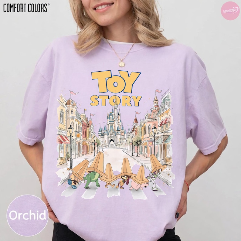 May include: Orchid-colored Comfort Colors t-shirt featuring a graphic from Toy Story. The design includes the words "Toy Story" in yellow, with a cartoon illustration of the characters crossing a street in front of a castle.