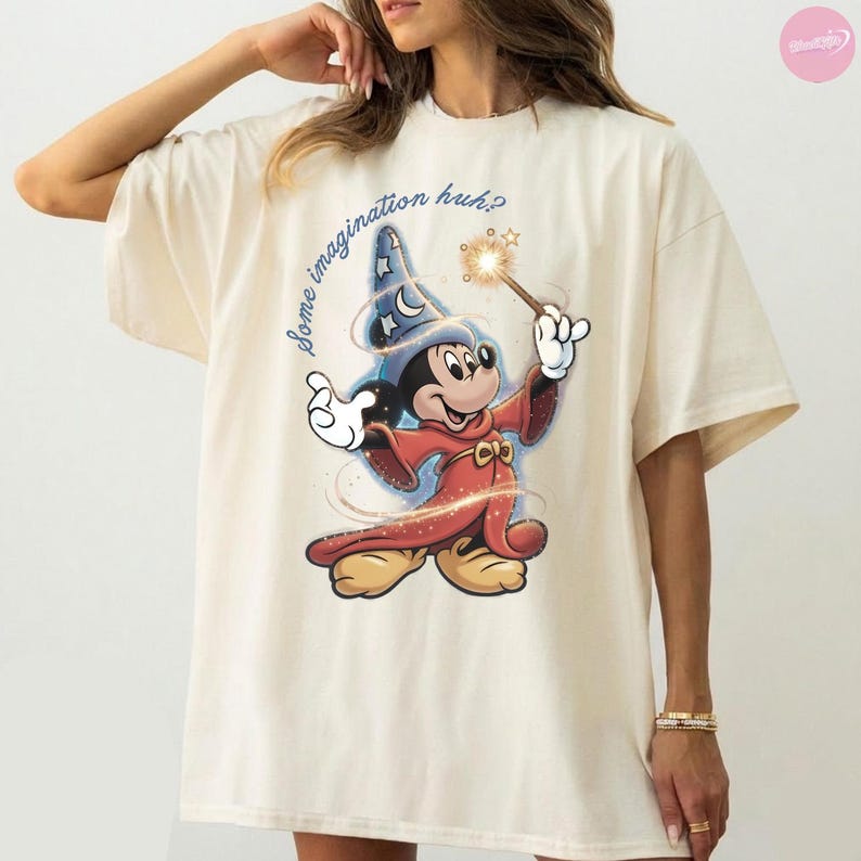 K&ouml;nnte beinhalten: Cremefarbenes T-Shirt mit einer Grafik von Mickey Mouse als Zauberer, der einen Zauberstab mit einem funkelnden Stern h&auml;lt. Der Text "some imagination huh?" steht in einer verspielten Schrift &uuml;ber der Figur. Das Design ist in Rot-, Blau- und Goldt&ouml;nen gehalten.