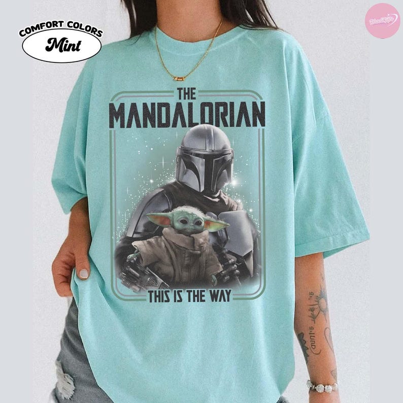 K&ouml;nnte beinhalten: Mintgr&uuml;nes T-Shirt mit einem Grafikdesign von "The Mandalorian". Das Design zeigt den Text "THE MANDALORIAN" &uuml;ber einem Bild des Mandalorianers und Grogu. Unter dem Bild steht der Text "THIS IS THE WAY".