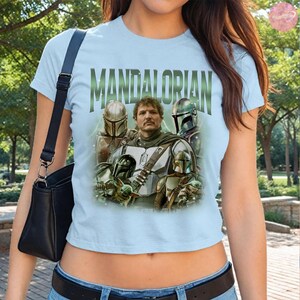 May include: Light blue t-shirt featuring the word "MANDALORIAN" in green block letters. The shirt displays a graphic of the Mandalorian and other characters from the series. The shirt is short-sleeved.