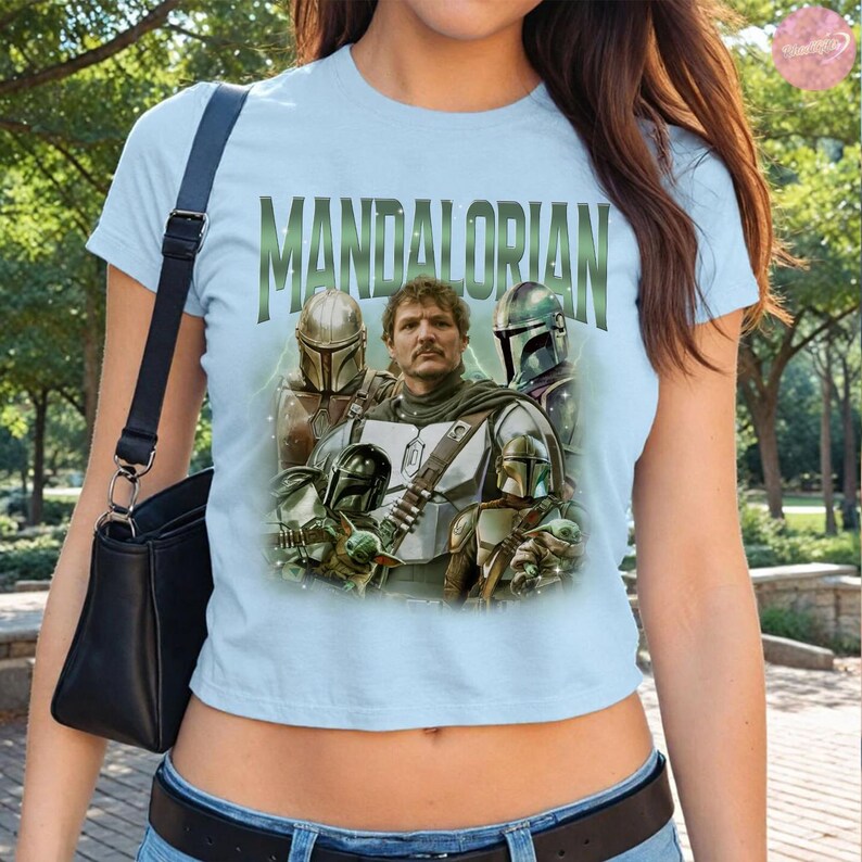 May include: Light blue t-shirt featuring the word "MANDALORIAN" in green block letters. The shirt displays a graphic of the Mandalorian and other characters from the series. The shirt is short-sleeved.
