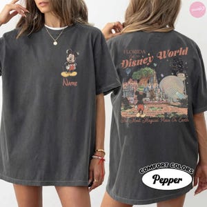 Puede incluir: Camiseta gris oscuro con Mickey Mouse en la parte delantera y la palabra "Name" debajo. La parte trasera muestra "Florida Disney World" con una escena del parque y "The Most Magical Place On Earth". La camiseta est&aacute; etiquetada como "Comfort Colors Pepper".
