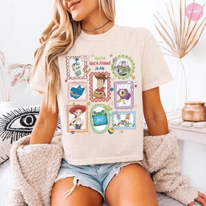 May include: A cream-colored t-shirt featuring a graphic print of various Toy Story characters arranged in postage stamp-style frames. The central text reads "You've Got A Friend In Me" surrounded by illustrations of Woody, Buzz Lightyear, Jessie, Rex, and other characters.