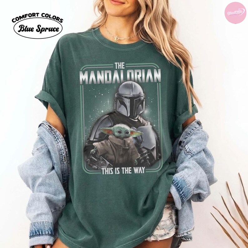 K&ouml;nnte beinhalten: Dunkelgr&uuml;nes T-Shirt mit einer Grafik von The Mandalorian und Grogu. Das Design enth&auml;lt den Text "THE MANDALORIAN" und "THIS IS THE WAY". Das T-Shirt ist von Comfort Colors und Blue Spruce.