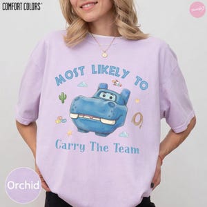 Op de afbeelding: Een lichtpaars T-shirt met korte mouwen en een blauwe cartoon nijlpaardafbeelding. De tekst luidt Most Likely To Carry The Team. Kleine illustraties van een cactus, raket, sterren en een lasso omringen het centrale nijlpaardfiguur.