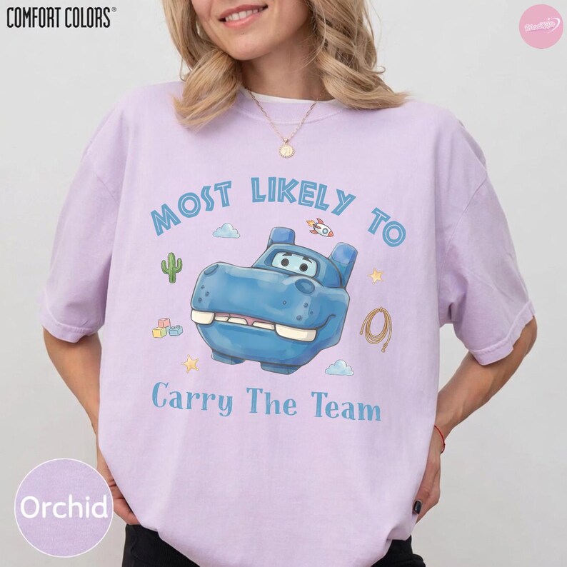 Op de afbeelding: Een lichtpaars T-shirt met korte mouwen en een blauwe cartoon nijlpaardafbeelding. De tekst luidt Most Likely To Carry The Team. Kleine illustraties van een cactus, raket, sterren en een lasso omringen het centrale nijlpaardfiguur.
