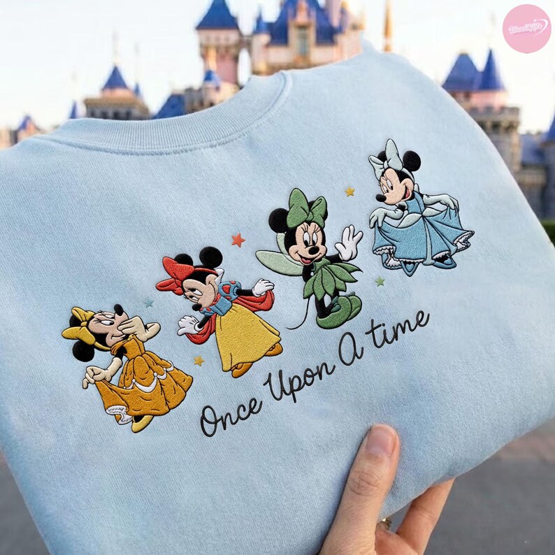 K&ouml;nnte beinhalten: Hellblaues Sweatshirt mit gestickten Minnie Mouse-Figuren in Prinzessinnen-Outfits. Der Schriftzug "Once Upon a time" ist in schwarzer Schrift gehalten. Das Sweatshirt wird von einer Person gehalten.