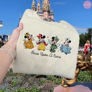 K&ouml;nnte beinhalten: Cremefarbenes Sweatshirt mit vier Minnie-Maus-Charakteren in Prinzessinnen-Outfits. Der Satz "Once Upon A Time" ist unter den Charakteren aufgedruckt. Der Hintergrund zeigt ein Schloss und einen blauen Himmel.
