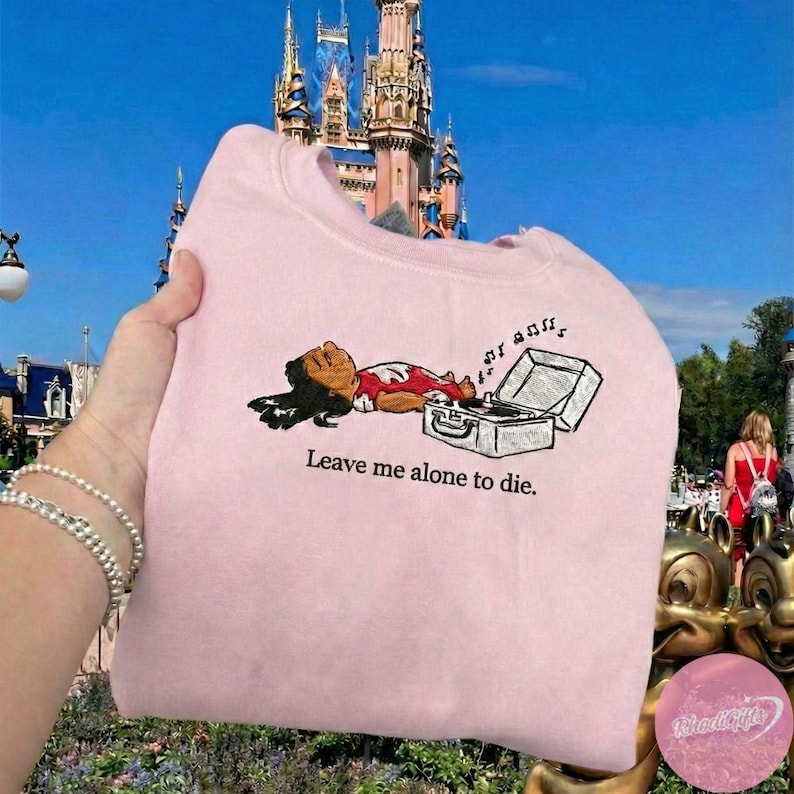 May include: A light pink sweatshirt with an embroidered graphic of a cartoon character lying next to a record player. The text "Leave me alone to die" is below the graphic. The background features a castle.