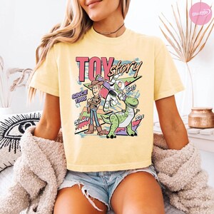 Op de afbeelding: Lichtgele t-shirt met een afbeelding van Toy Story-personages. De afbeelding bevat Woody, Buzz Lightyear en Rex, met de woorden "Toy Story" en "Since 1995". Het shirt heeft korte mouwen en een ronde hals.