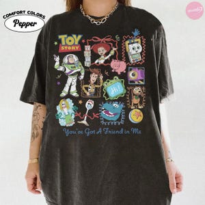 May include: A charcoal gray short-sleeve t-shirt featuring a graphic print of Toy Story characters including Buzz Lightyear, Woody, Jessie, and Forky. The design includes colorful illustrations of various movie icons and the text You've Got A Friend in Me.