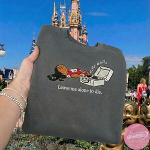 May include: Dark gray sweatshirt with an embroidered design of a person lying next to a record player, with musical notes above it. The text "Leave me alone to die." is below the design. The background features a castle.