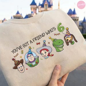 K&ouml;nnte beinhalten: Beiges Sweatshirt mit dem Schriftzug "YOU'VE GOT A FRIEND WHO'S 6" und Cartoonfiguren mit Partyh&uuml;ten. Das Design zeigt Woody, Buzz Lightyear, Jessie, Rex und Slinky Dog, mit einem gr&uuml;nen Ballon.