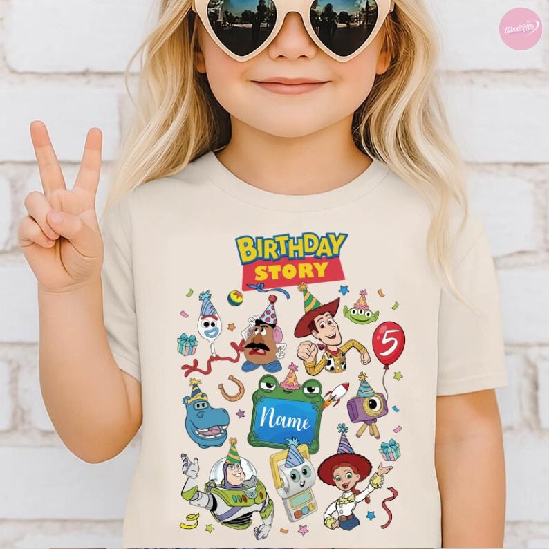 May include: A cream-colored t-shirt featuring a colorful graphic with the text Birthday Story. The design includes various cartoon characters wearing party hats, a red balloon with the number 5, and a green frame with the word Name inside.